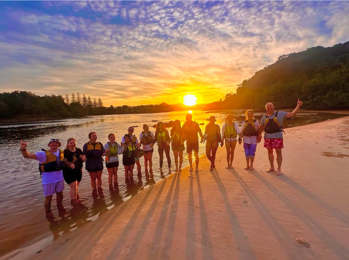 A tour group with paddles and lifejackets on the sand at the Brunswick River at sunset